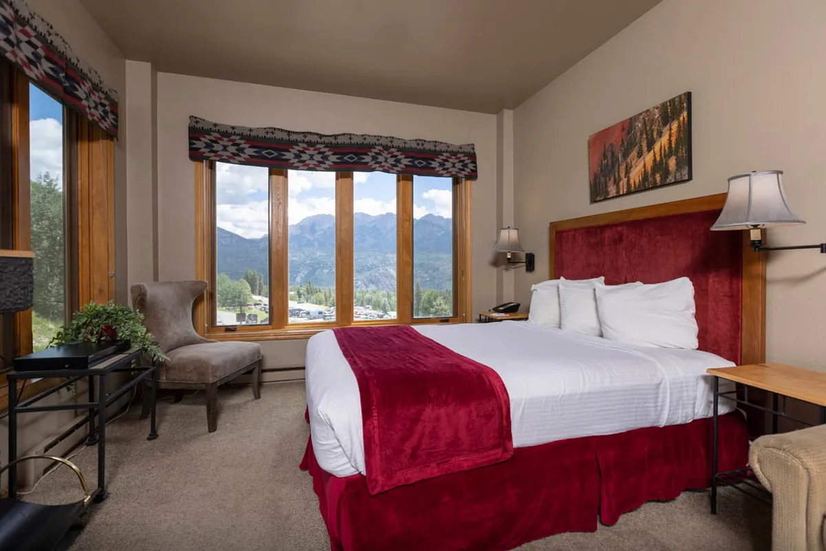 A serene bedroom with a plush bed featuring a red accent, large windows framing stunning mountain views, and a comfortable chair positioned to take in the scenery. Soft lighting and tranquil decor create an inviting, restful atmosphere.