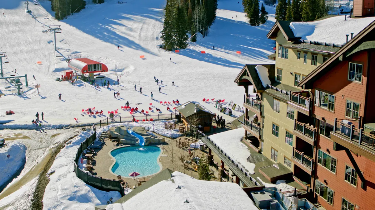 A vibrant exterior view featuring the ski slopes, a sparkling swimming pool, a ski lift, and the main lodge, all set against the backdrop of a scenic mountain landscape. The late afternoon light hints at après-ski gatherings and relaxation by the pool after an adventurous day on the slopes.