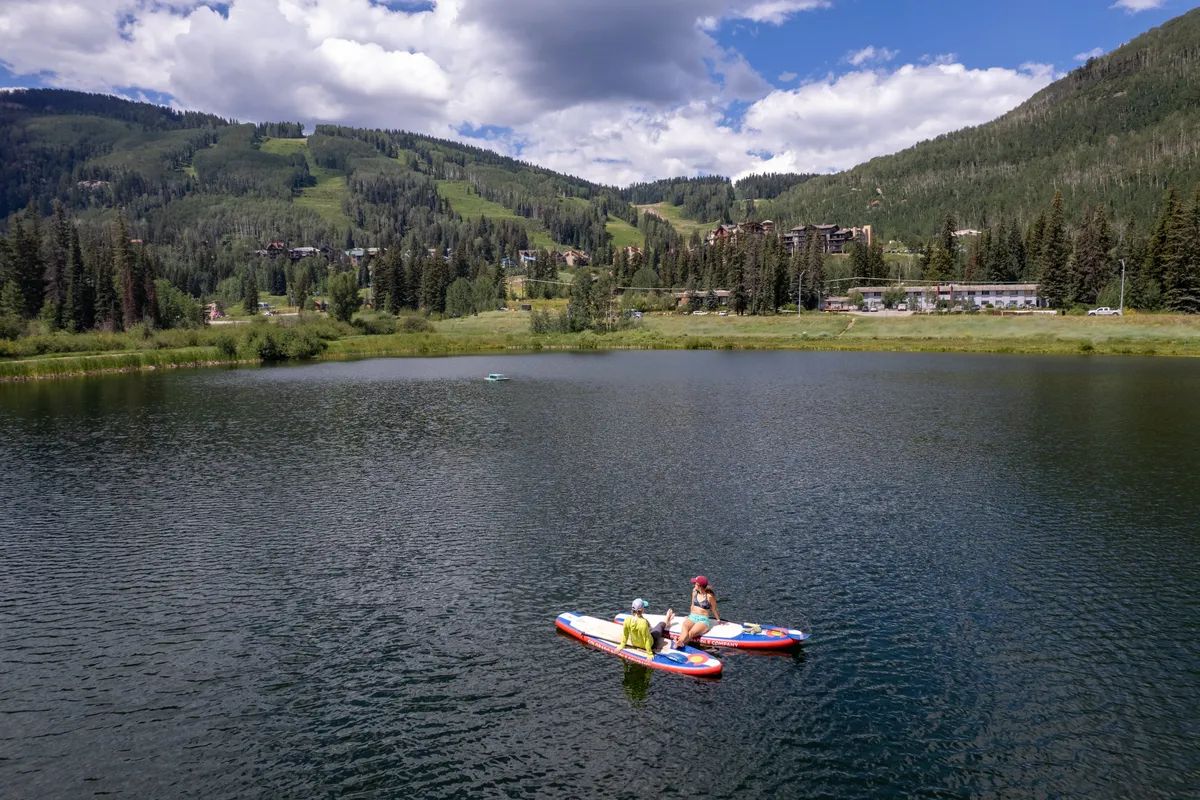 A serene summer setting with two people paddleboarding on a calm lake, surrounded by mountains. The scene conveys a sense of adventure, friendship, and the variety of outdoor activities available beyond winter sports.