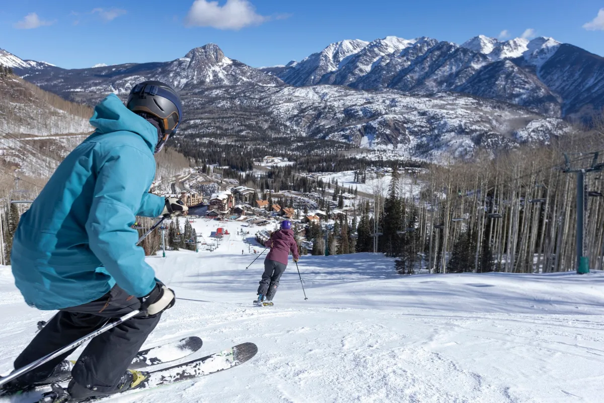 A dynamic scene showing a skier in the foreground carving through fresh snow, with another skier in the background, all set against majestic mountains and the lively ski resort village. The snow-covered slopes and bustling atmosphere evoke the excitement of winter sports.