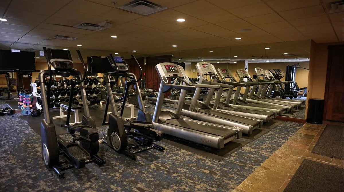 A modern interior fitness center with treadmills, ellipticals, dumbbells, and mirrored walls. The room is spacious and well-lit, with several guests engaged in workouts and group fitness classes, exuding a motivating and energetic atmosphere.