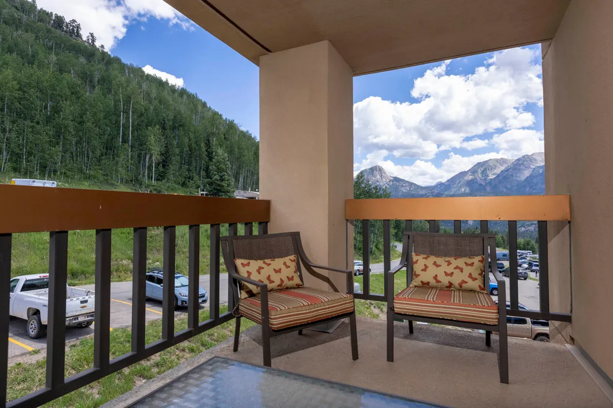 A tranquil balcony setting with comfortable seating and a glass table, looking out over breathtaking mountain vistas. The image captures the warm glow of sunset, inviting guests to relax and soak in the scenery together.