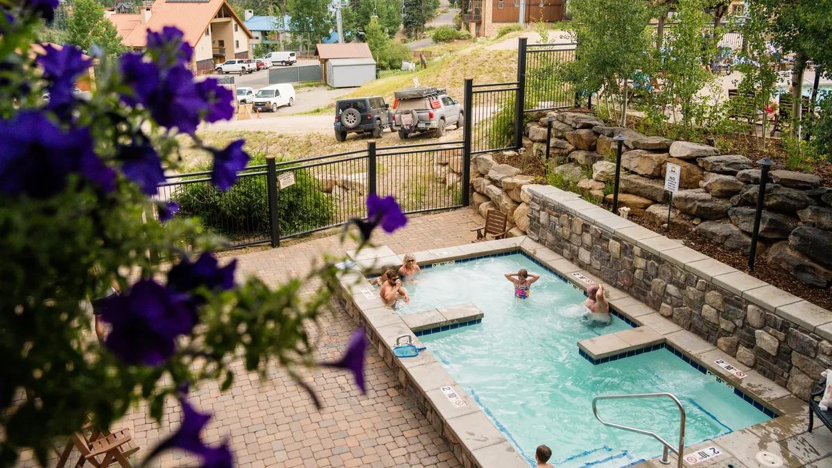 An exterior view of a heated outdoor swimming pool and adjacent hot tub, surrounded by lush flowers. People of all ages are enjoying the pool and hot tub, some swimming, others relaxing or socializing poolside. The atmosphere is bright and leisurely, emphasizing wellness and recreation.